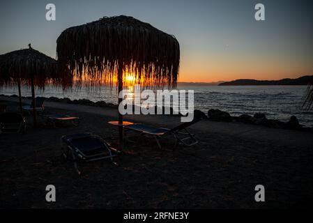 All'alba del resort, i raggi del sole sgorgano attraverso l'ombrellone dalle foglie di palma. Sagome di sdraio, ombrelloni e molo. Foto Stock
