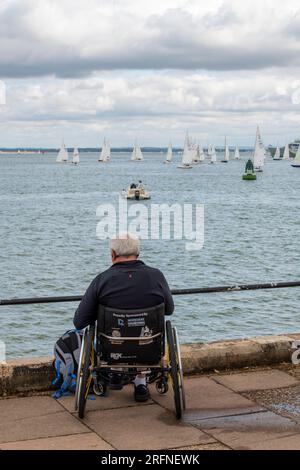 uomo seduto su una sedia a rotelle in riva al mare a guardare lo yacht che regtta la settimana annuale di cowes sull'isola di wight nel regno unito Foto Stock