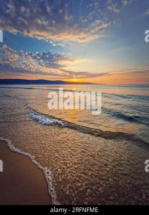 Vivace alba sul mare con onde schiumose che lavano la spiaggia di sabbia. Scena dell'alba con il sole che sorge sulle colline all'orizzonte Foto Stock