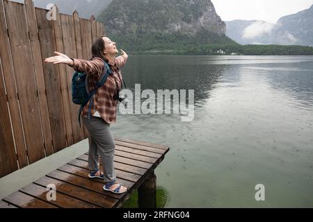 Giovane donna con una macchina fotografica in piedi su un palco di legno con le braccia spalancate e sorridenti, Hallstatt, Austria Foto Stock