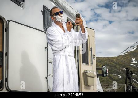 Uomo caucasico che si rasa il viso davanti al camper Van utilizzando la fotocamera dello smartphone Foto Stock