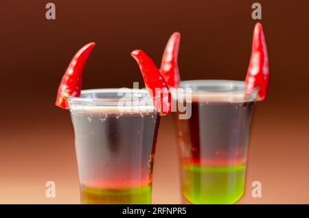 Shot di bevanda a strati preparato con vodka, liquore verde, granadina e succo di limone decorato con peperoncini a forma di corna, shot di diavolo Foto Stock