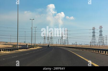 Un'immagine di un'autostrada in Kuwait, con più linee elettriche che la attraversano. Sullo sfondo sono visibili i camini con il fumo che esce da loro. Foto Stock