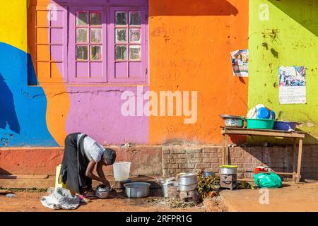 Di fronte ad una facciata dipinta di colori luminosi, una casalinga lava i piatti all'esterno. Jinja, Uganda. Foto Stock