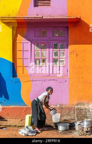 Di fronte ad una facciata dipinta di colori luminosi, una casalinga lava i piatti all'esterno. Jinja, Uganda. Foto Stock