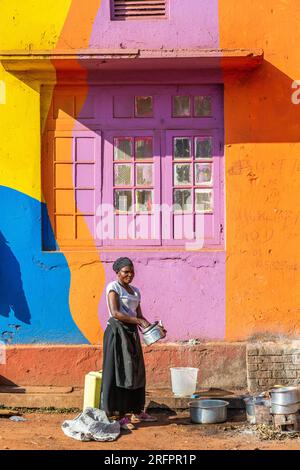 Di fronte ad una facciata dipinta di colori luminosi, una casalinga lava i piatti all'esterno. Jinja, Uganda. Foto Stock