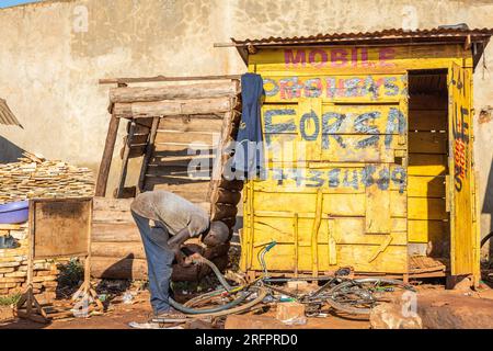 Di fronte a una stalla di legno, dipinta di giallo, un uomo sta riparando uno pneumatico per biciclette. Jinja, Uganda. Foto Stock