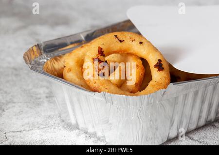 Anelli di cipolla in pastella in un contenitore di cartone con vassoio di alluminio da asporto. Su uno sfondo concreto Foto Stock