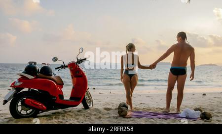 Gita su strada in scooter. Bella coppia in moto rossa in abiti bianchi sulla spiaggia di sabbia. Matrimonio appena sposato persone che camminano vicino a palme tropicali, mare. Luna di miele in riva al mare. Asia Thailandia. Noleggio moto. Foto Stock