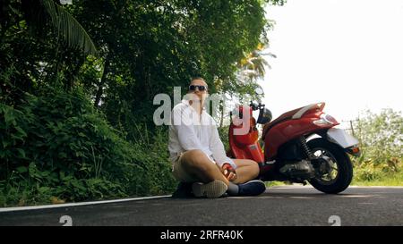 Un uomo in moto rossa in abiti bianchi guida lungo un sentiero nella foresta. Un uomo turista caucasico con occhiali da sole seduto sul marciapiede, rilassati, riposa, vicino allo scooter. Turismo in Asia e Thailandia. Noleggio moto. Foto Stock