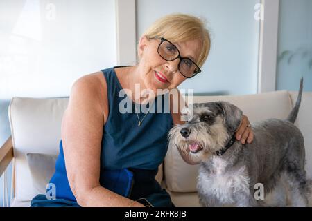 Allegra anziana anziana in pensione con le rughe che sorridono mentre abbraccia il suo cane Schnauzer e si diverte con il suo animale domestico a casa Foto Stock
