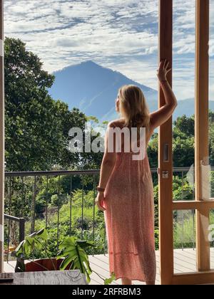 Una donna di mezza età in abito rosso si trova vicino alla porta del balcone e guarda la giungla e le montagne in una soleggiata giornata estiva Foto Stock