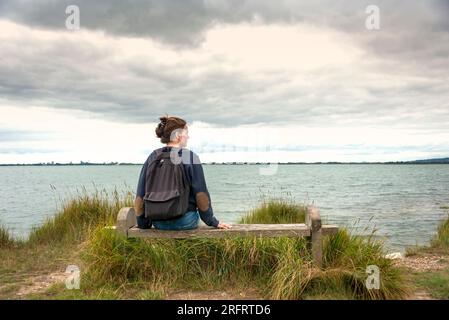 Donna solitaria seduta su una panchina vicino all'acqua, godendo la vista. Foto Stock