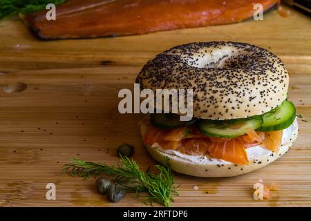 Bagel con salmone affumicato, formaggio cremoso, cetriolo, aneto e capperi su tavola di legno, spazio paesaggistico a sinistra. Foto Stock