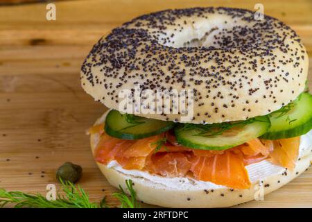 Bagel con semi di salmone affumicato, formaggio cremoso, cetriolo, aneto e capperi su tavola di legno, paesaggio. Foto Stock