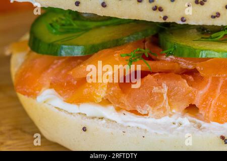 Bagel con semi di salmone affumicato, formaggio cremoso, cetriolo, aneto e capperi su tavola di legno, paesaggio, macro. Foto Stock