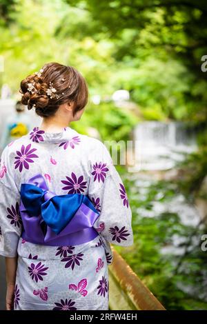 Vista posteriore di una donna che indossa un kimono estivo yukata giapponese che cammina lungo la strada accanto alla cascata della foresta in natura. Kyoto, Giappone. Foto Stock