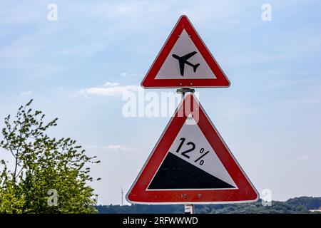 Segnali stradali indicanti: Segnale di avvertimento aeroportuale o aereo a bassa quota e pendenza ripida del 12% in discesa su strada, contro il cielo blu su una somma soleggiata Foto Stock