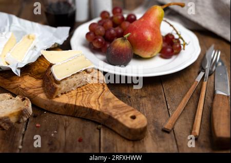 Affettare il formaggio Camembert su un pezzo di pane con frutta e un bicchiere di vino sullo sfondo Foto Stock