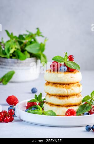 Pancake spessi con formaggio cottage con mirtilli, lamponi e ribes rosso, decorati con menta su piatto bianco, colazione deliziosa. Tavola grigia Foto Stock