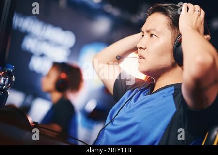 Vista laterale di un ragazzo asiatico confuso, un giocatore di cyber sport maschile che indossa cuffie che guardano lo schermo del PC con una triste espressione facciale mentre partecipa Foto Stock