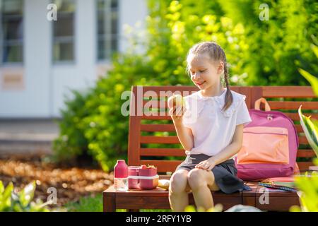 Allievo con pranzo al sacco in mano. La ragazza con gli zaini sta mangiando frutta vicino all'edificio all'aperto. Inizio delle lezioni. Primi giorni di autunno. Foto Stock