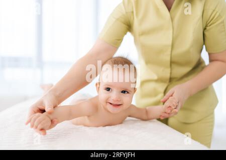 Foto corta di un massaggiatore pediatrico irriconoscibile con un bambino carino che fa esercizio fisico semplici esercizi per rendere più forti le ossa e i muscoli del bambino. Foto Stock
