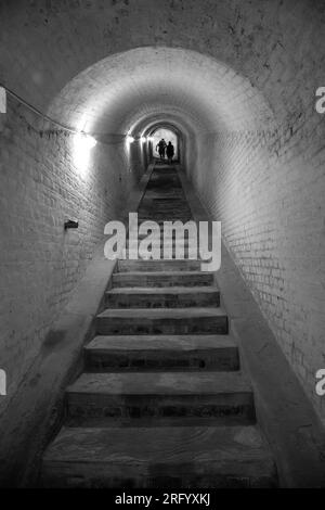 Foto monocromatica di scale in pietra all'interno di un tunnel al forte Drop Redoubt di dover, Inghilterra, con un gruppo di persone in cima. Foto Stock