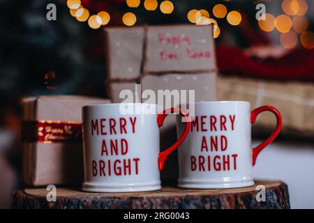 Due tazze bianche con testo natalizio rosso in piedi su un ceppo di legno. Decorazioni per le vacanze invernali, dettagli per Capodanno Foto Stock