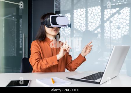 Donna asiatica che indossa occhiali per realtà virtuale seduta a una scrivania in un'area di lavoro all'interno dell'ufficio, donna d'affari che parla in remoto con i colleghi, simulazione di riunioni virtuali. Foto Stock