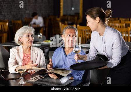 Cameriera che porta il vino alla coppia anziana scegliendo piatti dal menu Foto Stock