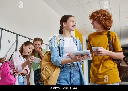 ragazzo e ragazza adolescente felici che tengono gadget vicino ai compagni di classe nel corridoio scolastico, concetto di ritorno a scuola Foto Stock