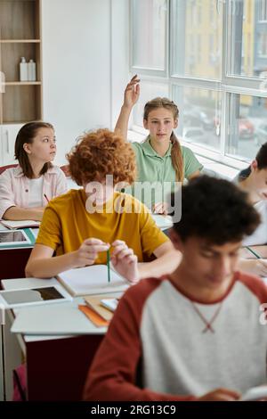 Studentessa adolescente che alza la mano e parla vicino a dispositivi e compagni di classe durante la lezione a scuola Foto Stock