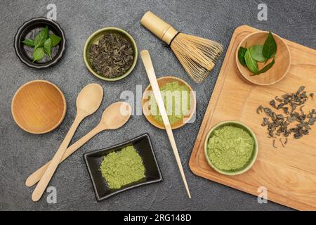 Foglie verdi, polvere di tè matcha su tagliere e tavolo. Frusta di bambù e cucchiaio sul tavolo. Spianatura piatta. Sfondo nero Foto Stock