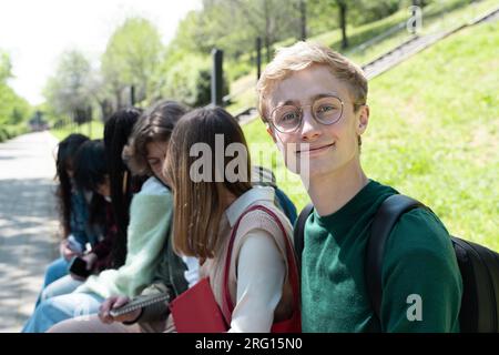 Un gruppo di adolescenti multirazziali siede in un parco. Mentre gli amici interagiscono tra loro, un ragazzo britannico con occhiali, capelli biondi e lentiggini Foto Stock