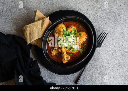 Vista dall'alto del piatto georgiano Chakhokhbili è la carne di pollo stufata in salsa di pomodoro con spezie ed erbe servite su piatto nero con un po' di pane su tavola grigia Foto Stock