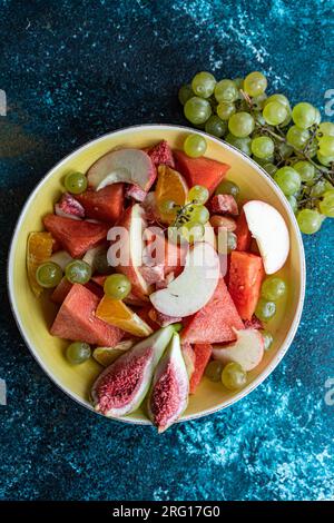 Vista dall'alto della ciotola in ceramica con fette di diversi tipi di frutta collocate sulla superficie blu vicino all'uva sfocata Foto Stock