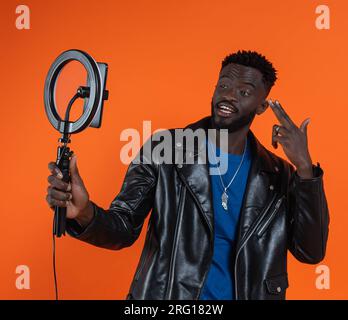 Happy African American maschio in giacca di pelle con smartphone e illuminatore anulare a LED con registrazione su supporto video su sfondo arancione delle riprese in studio Foto Stock