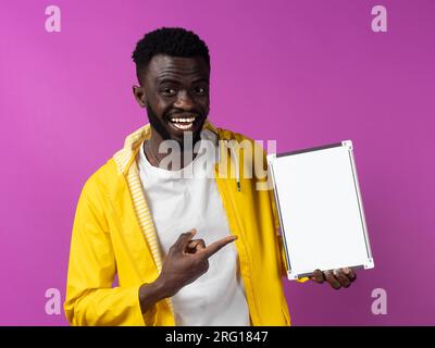 Un giovane sorridente in abiti casual con la bocca aperta che guarda la fotocamera mentre si trova in piedi e punta la mini lavagna bianca in mano contro lo sfondo viola Foto Stock