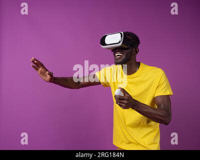 Eccitato giovane uomo afroamericano in abiti casual e cuffie per realtà virtuale mentre si è in piedi e si utilizza il controller con la mano e guardando gli interessi Foto Stock