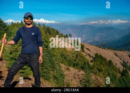 14 ottobre 2022, Uttarakhand India. Giovane escursionista solista con bastone da passeggio di bambù sulle colline himalayane, sfondo di cime glaciali. Uttarakhand, Ind Foto Stock