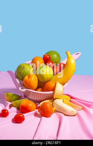 Recipiente di plastica con frutta fresca assortita, posto sul tavolo con un tavolo di tovaglia rosa alla luce del giorno su sfondo blu Foto Stock