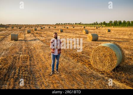 Agricoltore felice è in piedi accanto alle balle di fieno. È soddisfatto a causa del raccolto riuscito. Foto Stock