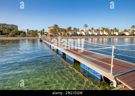 Ponte di ferro e passerella in legno sulla spiaggia cristallina di El Mar de Cristal, nel Mar Menor, Cartagena, Spagna, con la città sullo sfondo Foto Stock