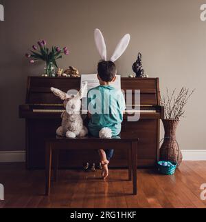 Ragazzo che indossa le orecchie conigliate che suona il pianoforte con il coniglio imbottito accanto a lui. Foto Stock