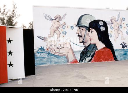 Bassora, Iraq 1984. Murale raffigurante la bandiera irachena con raffigurazione di Saddam Hussein che indossa l'uniforme dell'esercito accanto a una donna con fiori tra i capelli che reggono gioielli d'oro, fiancheggiato da cherubini e scena di sfondo di un carro armato e un cavaliere che carica con la spada estratta. 1980 HOMER SYKES Foto Stock