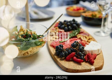 Tavola di legno con uva fresca e salame accanto alla ciotola di vetro con antipasto fatto in casa composto da fusilli e foglie di basilico sul tavolo servito Foto Stock