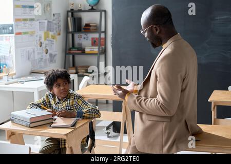 Insegnante afroamericano che spiega il nuovo argomento a scolaro mentre si siede alla scrivania e prende appunti, studiano dopo le lezioni Foto Stock