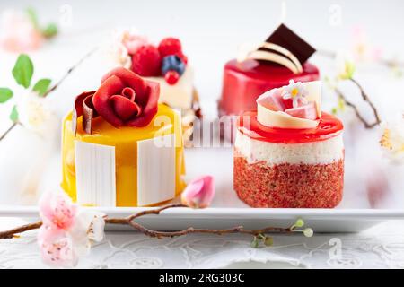 Varie mini torte su un piatto bianco. Dolci decorati con frutti di bosco freschi e fiori per le vacanze. Foto Stock