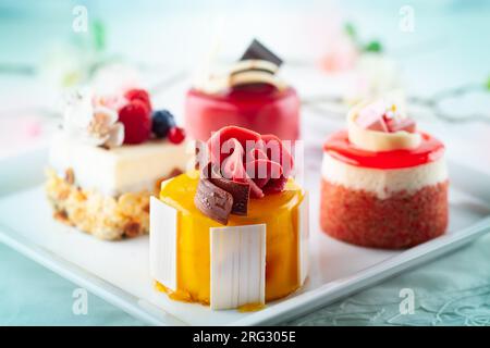 Varie mini torte su un piatto bianco. Dolci decorati con frutti di bosco freschi e fiori per le vacanze. Foto Stock